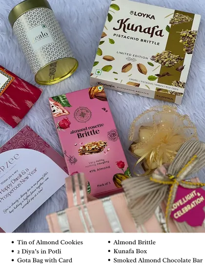 A flat lay of the Gota Bag Hamper contents. This Diwali gift includes almond cookies, almond brittle, a Kunafa box, smoked almond chocolate, and two diyas in a small potli.