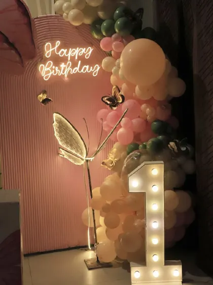 A close-up of a first birthday setup, highlighting the neon "Happy Birthday" sign, a large marquee '1', and a glowing butterfly prop.