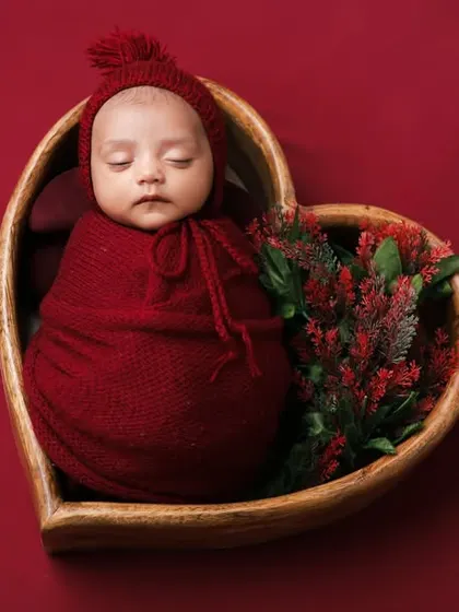 A newborn swaddled in red, nestled inside a heart-shaped wooden bowl. This is a perfect setup to symbolize the love for your new arrival.