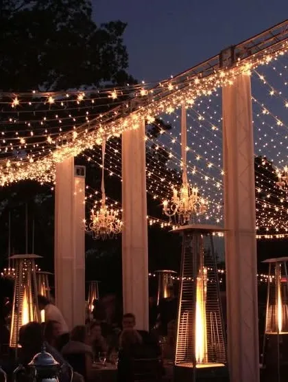 A magical evening setup for an outdoor event. The space is illuminated by a ceiling of fairy lights and multiple chandeliers, with patio heaters providing warmth and ambiance.