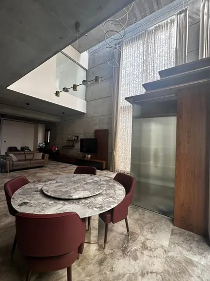 This open-plan dining and living area in a concrete-finished home showcases a double-height ceiling and expansive windows, creating a grand and airy atmosphere.