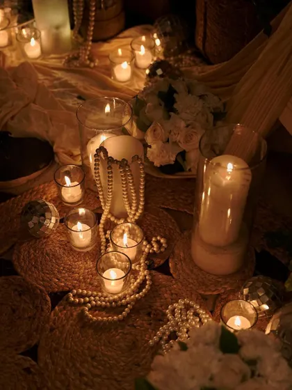 A close-up of the candlelit decor, showing pillar candles in glass hurricanes, strings of pearls, and mini disco balls scattered on rustic jute mats. This mix of textures creates a chic and eclectic look.