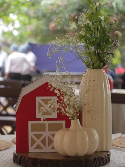 A simple yet charming centerpiece with a miniature red barn, white ceramic vases with baby's breath, and a white pumpkin, all set on a rustic wood slice.