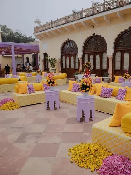 Guest seating for a Haldi ceremony in a palace courtyard. Low seating with yellow and purple cushions creates a comfortable and royal atmosphere.