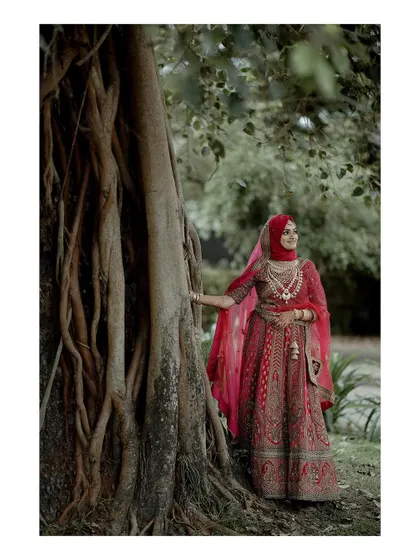 The bride stands gracefully by a majestic banyan tree, her red lehenga a vibrant splash of color against the natural backdrop.