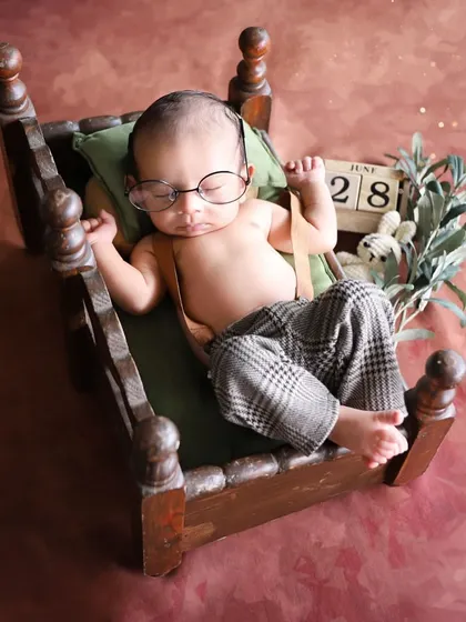 A full view of this charming, vintage-inspired setup. A baby wearing tiny glasses and suspenders sleeps on a miniature wooden bed, creating a quirky and adorable portrait.