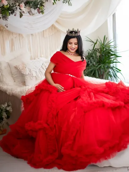 A queen on her throne. This mom-to-be is seated on our studio bed setup, draped in a magnificent red gown and wearing a crown, creating a portrait of pure elegance.