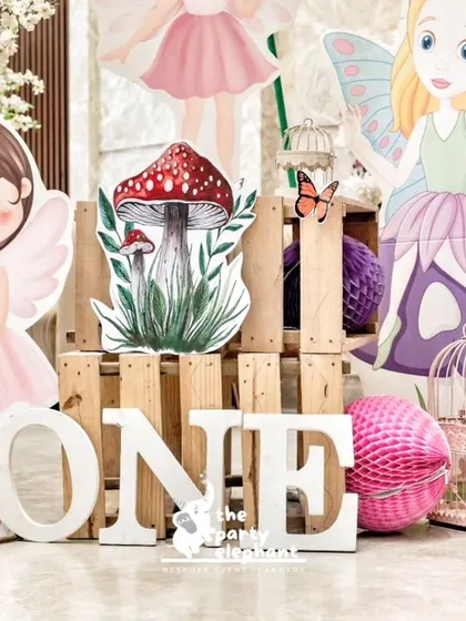 A charming display for a first birthday, with a "ONE" sign surrounded by adorable fairy cutouts and oversized mushroom props.