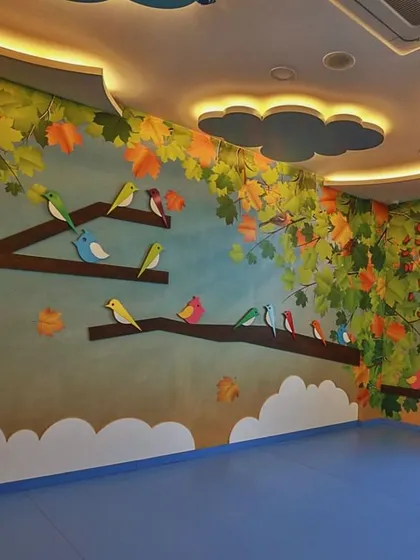 This classroom is inspired by a forest canopy, with colorful birds perched on tree branches painted on the walls. The ceiling features cloud-shaped light fixtures, bringing the feeling of being outdoors inside.