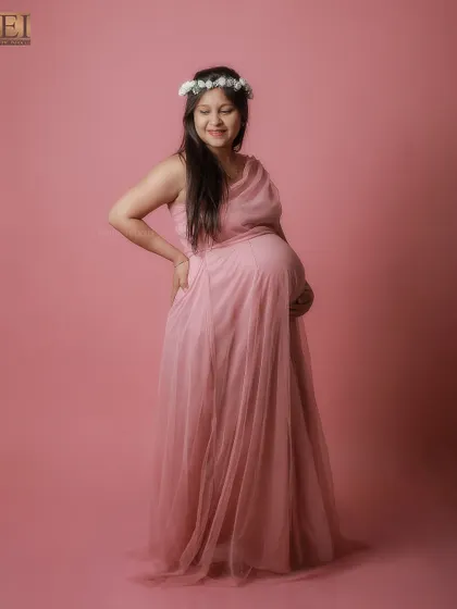 A smiling portrait in a pink gown and a delicate flower crown, set against a matching pink studio background.