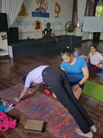 A teacher provides a hands-on adjustment to help a student lift their hips and lengthen their spine in Downward-Facing Dog.