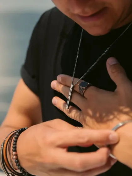 A man adjusting his silver-toned bar pendant, shown here with other accessories like rings and bracelets. This highlights the pendant as part of a complete, styled look.