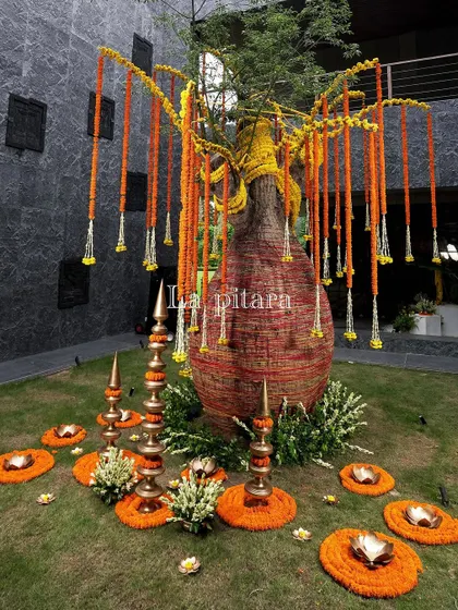 An outdoor art installation featuring a large decorative urn with marigold garlands hanging like a willow tree. This creative piece, surrounded by brass lamps and floral rangoli, blended tradition with artistic expression.