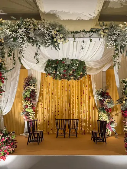 An intimate and traditional wedding mandap, rich with florals in hues of red, yellow, and white, and a backdrop of delicate hanging rose strings.
