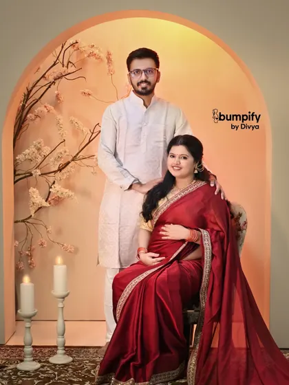 A classic seated portrait during a maternity session. The expecting mother, radiant in her red saree, is joined by her partner in our warm, arched studio setup.