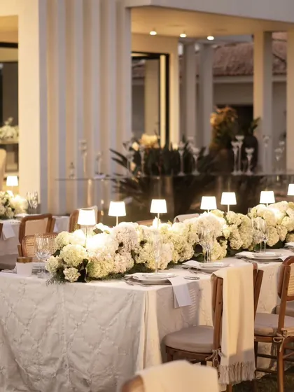 The warm glow of table lamps illuminates a long dining table set against the backdrop of a modern building. The contrast between the soft decor and the clean architecture is beautiful.