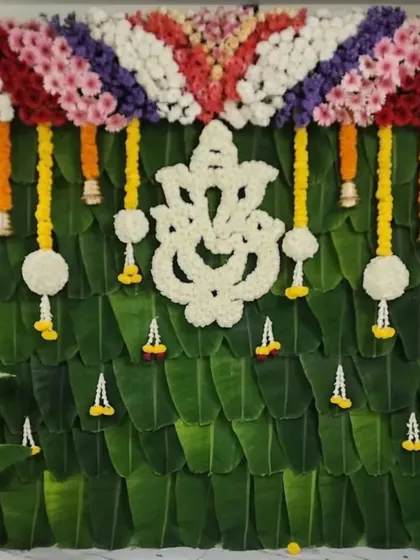 A traditional backdrop for a home ceremony, featuring a Ganesha made of white flowers against a wall of banana leaves. The colorful floral border adds a festive touch.