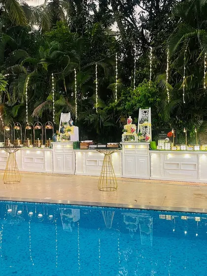 A stunning poolside catering setup for a milestone celebration. The clean, white buffet counters are beautifully reflected in the water, with fairy lights adding a touch of magic to the evening.