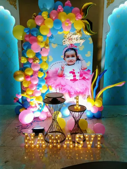 A colorful and personalized first birthday decoration. This setup includes a custom backdrop with the baby's photo, a vibrant balloon arch, and light-up marquee letters for the name.