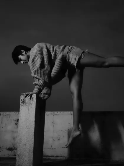 A striking black and white photo capturing a model in an athletic, dance-like pose on a concrete ledge. The image highlights form and movement against a stark, dark sky.