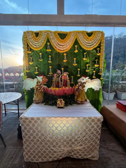 A sacred corner dedicated to tradition. This setup features deities on a lotus base, set against a backdrop of fresh banana leaves and garlands of marigold and tuberose flowers.