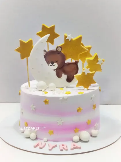 A dreamy cake设计 featuring a teddy bear sleeping on the moon. The soft pink and white ombre and golden stars make it perfect for a baby's celebration.