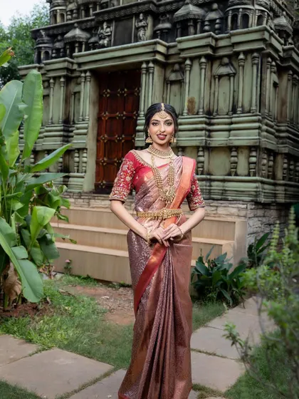 A bride in a beautiful bronze and red silk saree, standing before a temple. This earthy and warm color combination is perfect for a traditional and elegant look.
