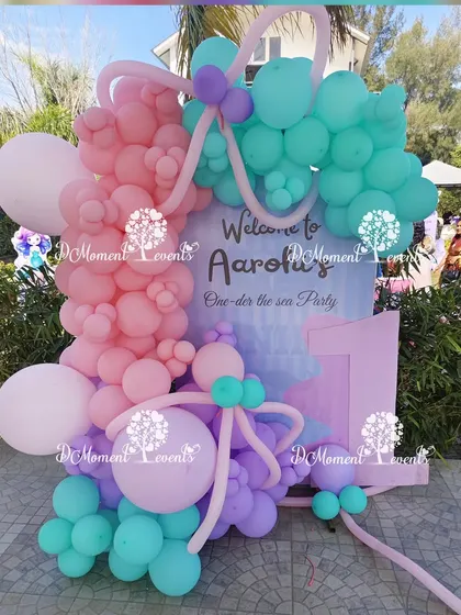 A welcome sign and backdrop for a "One-der the Sea" first birthday, decorated with a beautiful arrangement of pink, purple, and teal balloons.
