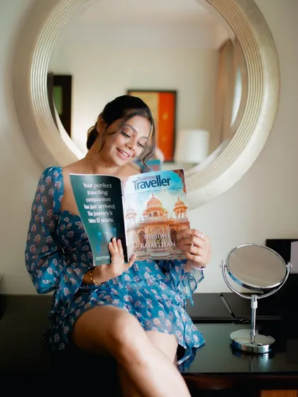 A lifestyle shot capturing a relaxed, indoor moment. The model is reading a travel magazine, creating a narrative that connects the fashion (a floral dress) with a sophisticated, leisurely lifestyle.