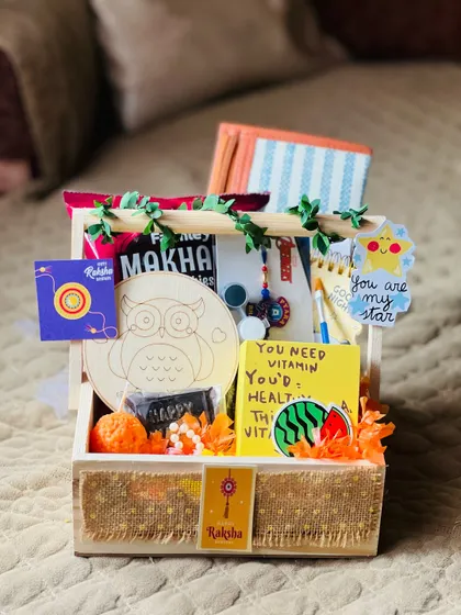 A creative Rakhi hamper for kids, including a DIY owl painting kit, a notepad, and other fun items in a jute-lined crate.