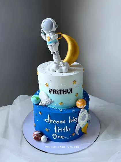 "Dream big little one" with this space-themed cake. This two-tiered design features an astronaut sitting on the moon, surrounded by planets and a rocket ship.