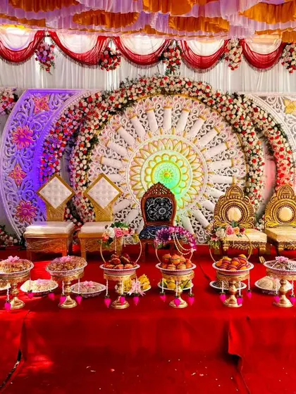 This is a highly ornate and traditional stage for a wedding ceremony. It features intricate white and gold circular backdrops, multiple thrones, and a table laden with decorative trays filled with sweets and offerings.