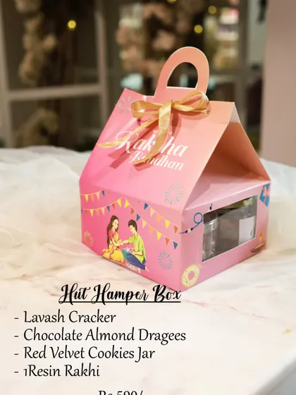 The Hut Hamper Box for Raksha Bandhan is a cute and compact gift. It contains a pack of lavash crackers, chocolate almond dragees, a jar of red velvet cookies, and a resin rakhi.