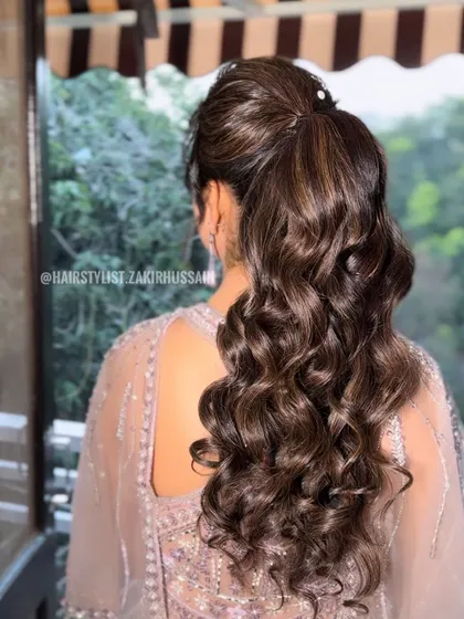 A glamorous high ponytail with voluminous curls, perfect for a reception or cocktail party. The hair is wrapped at the base for a sleek finish, and the highlights add beautiful dimension to the curls.