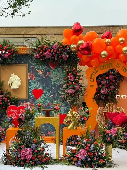 The complete "A Little Cutie Is On The Way" baby shower setup, featuring a lush floral and balloon arch against a tropical print backdrop. The design incorporates a lit-up heart, a peacock chair for the mom-to-be, and a vibrant orange, red, and gold color scheme.