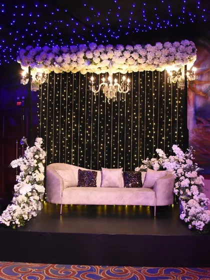 A cozy and glamorous seating area for a Sangeet. A plush couch is set against a backdrop of twinkling fairy lights, with a canopy of white flowers and a chandelier adding to the starry night ambiance.