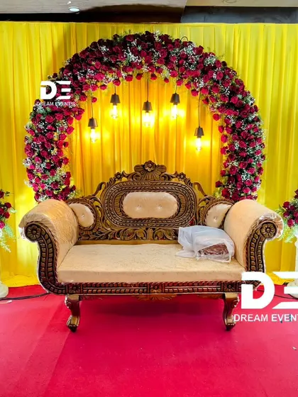 A simple yet striking engagement stage with a bold yellow backdrop. The circular arch of red roses and hanging Edison bulbs provides a beautiful contrast, making the ornate sofa stand out.