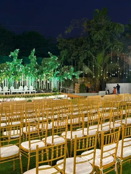 A wide view of the enchanted forest reception setup. The golden guest chairs lead the eye towards the magical stage, with fairy lights twinkling in the trees above, creating a cohesive and immersive environment.