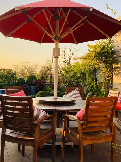 A beautiful shot of the terrace dining area at sunset. The wooden table and chairs, red cushions, and lush surrounding plants create a warm and inviting scene.
