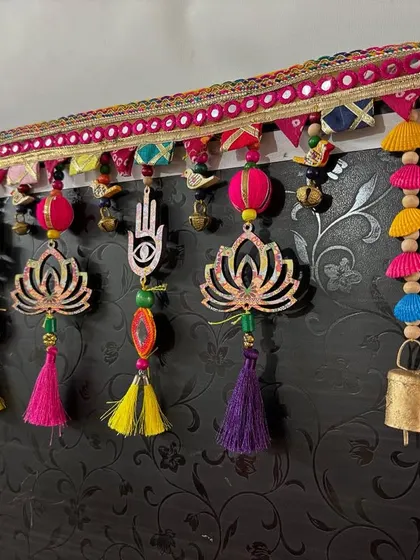 A close-up of the 'Shubh Toran' hanging against a dark door, which really makes the vibrant pinks, yellows, and purples pop. The mirror work on the topper adds a festive sparkle.
