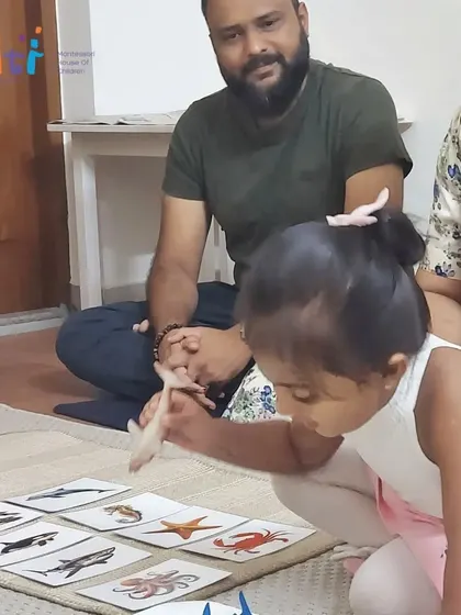A child shows her parents how she can match animal figures to picture cards. This activity builds vocabulary and classification skills, which are foundational for scientific understanding.
