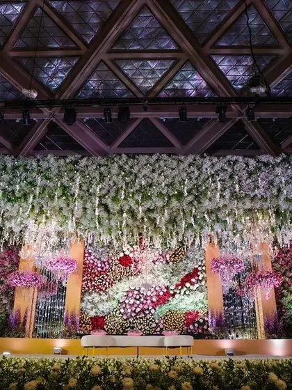 This stage design creates a "magical starry night" effect with a dense canopy of hanging white flowers and intricate lighting. The backdrop is a rich tapestry of red and white florals, demonstrating a deep and layered approach to stage decoration.