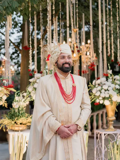 A smiling groom stands before a backdrop of hanging floral strings and chandeliers at his Lohagarh Fort wedding. The decor blends traditional elements with opulent touches.