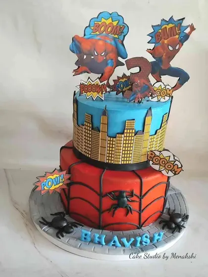 An action-packed two-tier Spider-Man cake. The bottom tier features a spiderweb design, while the top shows the city skyline, complete with fun "Boom!" and "Pow!" signs.
