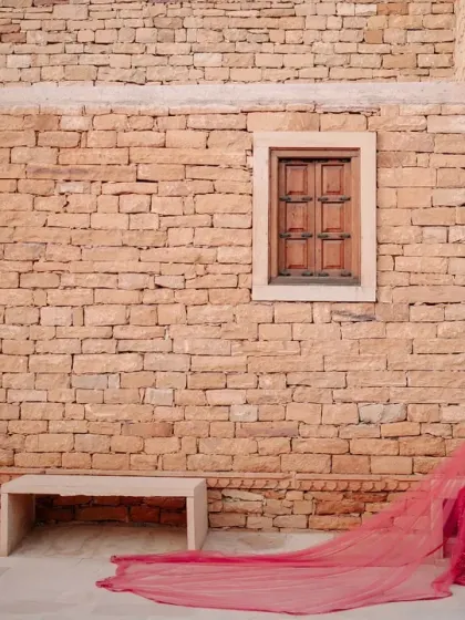 A breathtaking bridal portrait in Jaisalmer, with the bride's long red veil trailing behind her against a rustic stone wall.