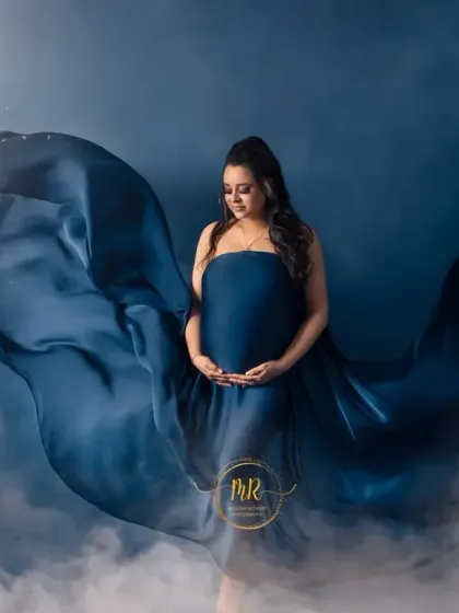 A celestial-themed maternity portrait. The mother-to-be is wrapped in flowing blue fabric against a night sky backdrop with a full moon, creating a serene and magical image.