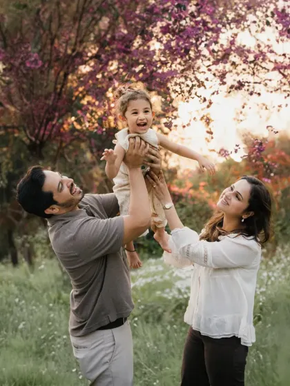 Parents lift their laughing daughter into the air against a backdrop of beautiful pink-flowered trees. This is a moment of pure, uninhibited family fun.