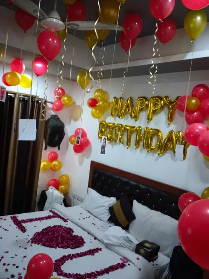 A romantic birthday setup with red and gold balloons, "Happy Birthday" foil letters, and "I ❤️ U" written with rose petals on the bed.