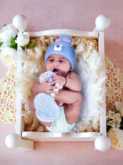 A cute baby boy in a little bed prop, looking curious and playful. The soft pink background and white floral accents create a gentle and charming setting for this infant portrait.