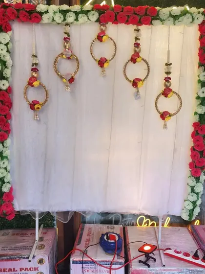 A simple and elegant backdrop for your Ganapati idol. This design uses a white curtain with a floral border and delicate hanging ornaments, all illuminated by warm fairy lights for a serene glow.